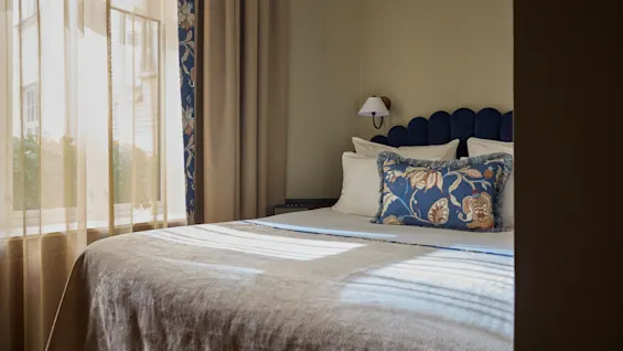 A neatly made bed with decorative pillows is positioned by a window with sheer curtains, allowing soft daylight to illuminate the room. A wall lamp adds ambient lighting.