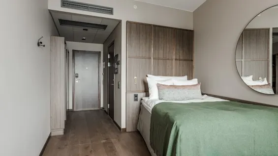 En pænt redt seng med hvidt og grønt sengetøj står ved siden af et rundt spejl på en beige væg i et moderne hotelværelse med trækaccent.