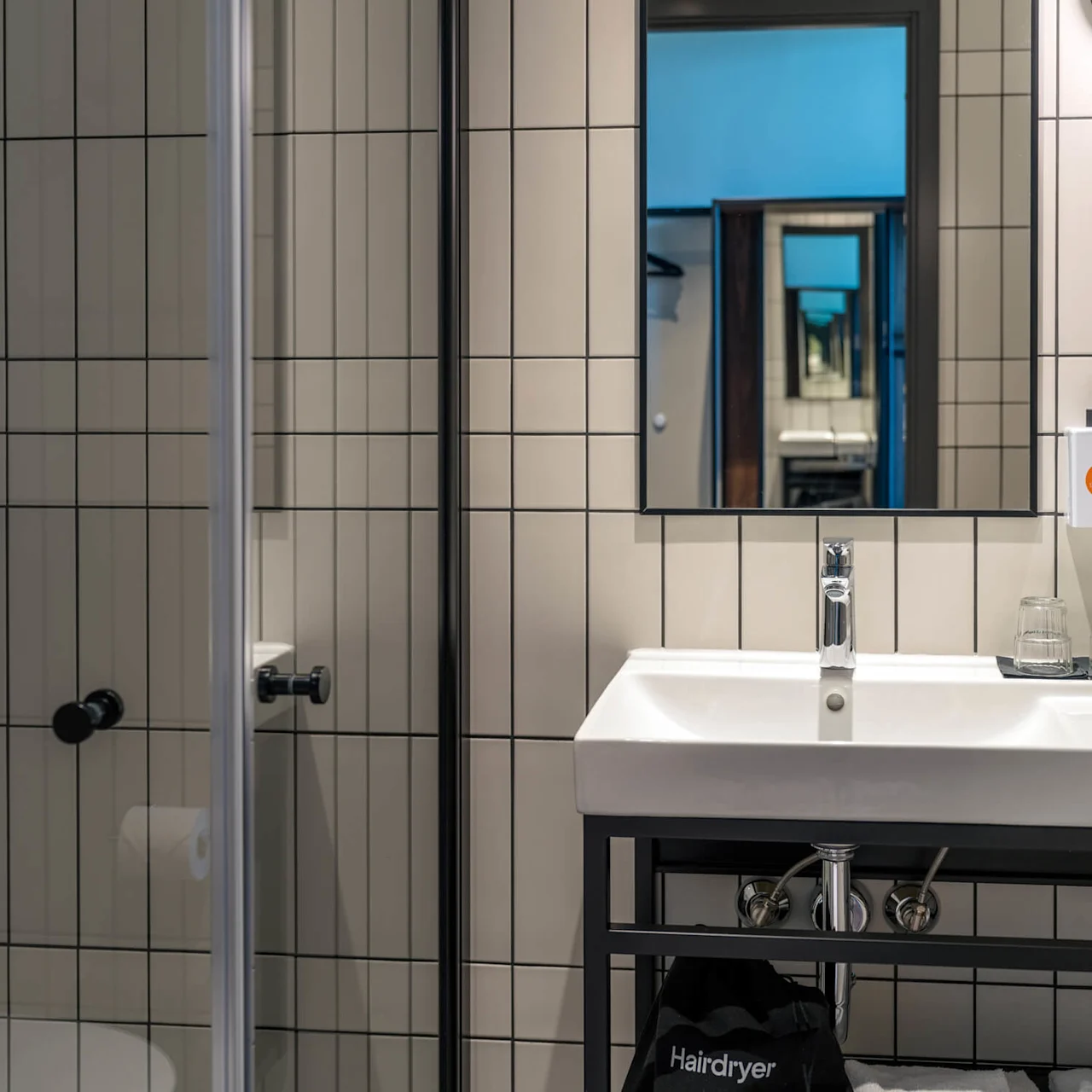 Dusj med metallarmatur og såpedispensere i flislagt bad. Ved siden av er en vask under et speil som reflekterer flere bilder. Tekst inkluderer "Hairdryer."