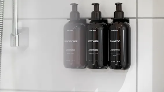 Tre mørke pumpeflasker merket balsam, kroppssåpe og sjampo montert på en hvitflislagt dusjvegg.