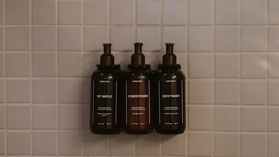 Tre mørke pumpeflasker mærket "SHAMPOO," "CONDITIONER," og "BODY WASH" er monteret mod en flisebelagt væg. Hver etiket læser: "BASICS" efterfulgt af mindre tekst.