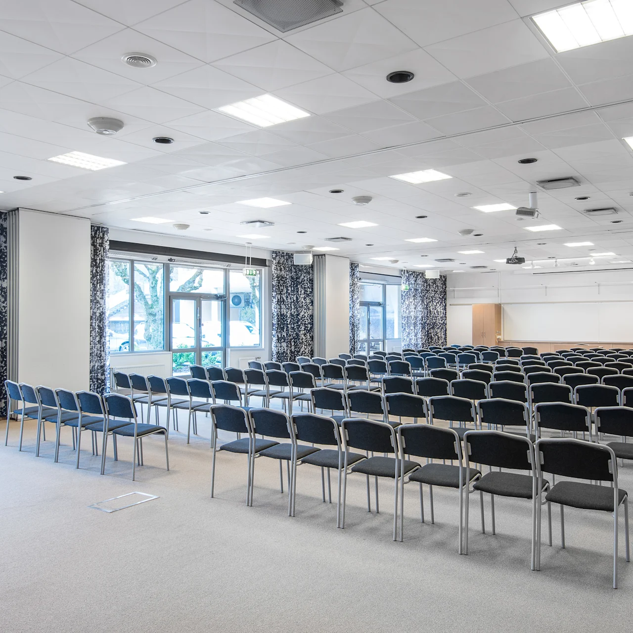 Rader med tomme svarte stoler vender fremover i et godt opplyst konferanserom med store vinduer dekket av mønstrede gardiner. Rommet er klargjort for et arrangement eller møte.