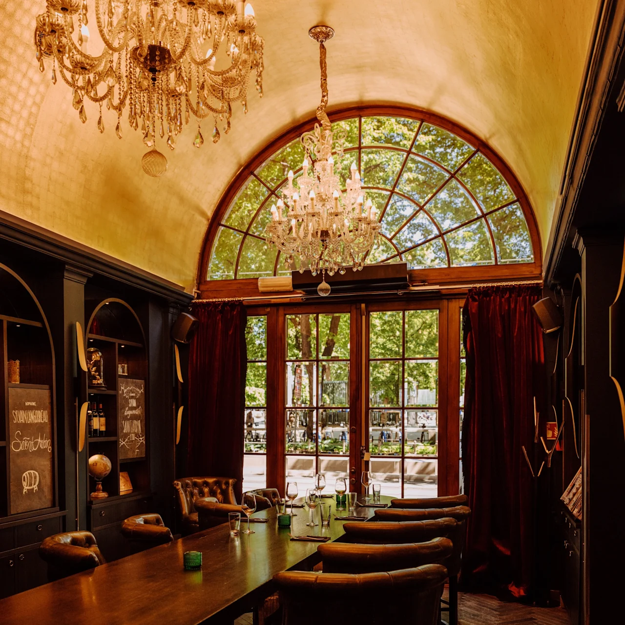 A long wooden table with chairs sits beneath ornate chandeliers in an elegant room, with a large arched window providing a view of leafy trees and sunlight streaming inside.