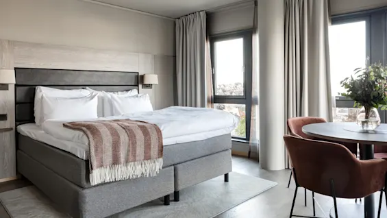 A neatly made bed with a striped throw lies beside a small round table with flowers. The room is bright with large windows and cream curtains.