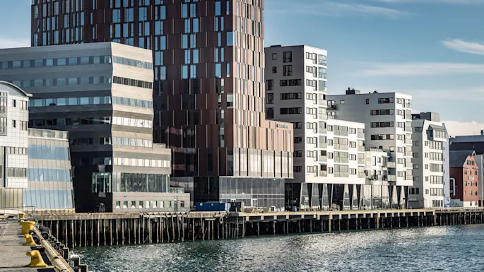 Modern buildings stand along a waterfront, reflecting sunlight. The largest structure is a brown, glass-panelled tower. The text "Radisson Blu" is visible on one building.