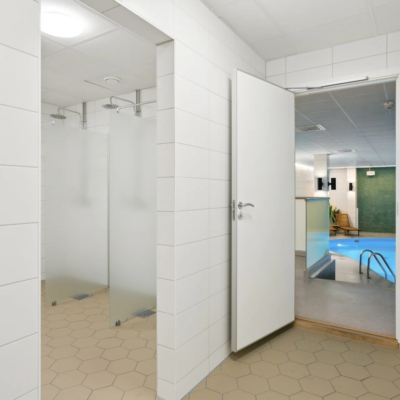 Bruseområde med matteret glas og flisegulv, ved siden af en åben dør der fører til en indendørs swimmingpool med blåt vand i en moderne facilitet.