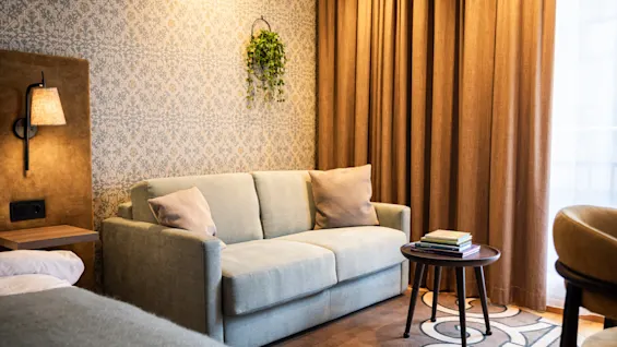 En beige soffa står med kuddar i ett mysigt hotellrum, bredvid en mönstrad tapetserad vägg och bruna gardiner. Ett träbord bredvid har en hög med böcker.