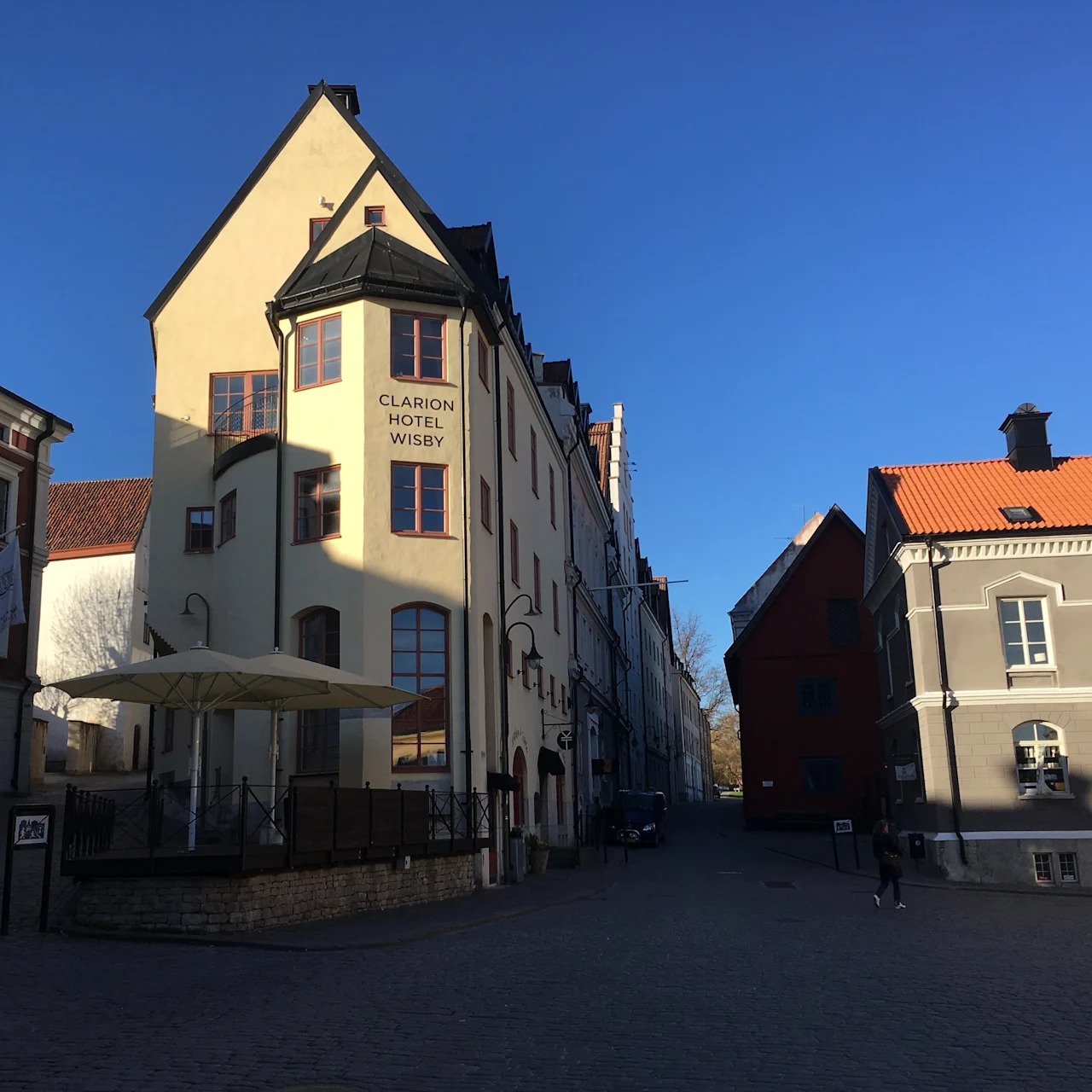 Clarion Hotel Wisby, med sin beige facade og høje buede vinduer, står på en brostensbelagt gade under en klar blå himmel. Nærliggende bygninger kaster skygger på jorden.