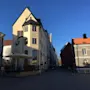 Clarion Hotel Wisby, med sin beige facade og høje buede vinduer, står på en brostensbelagt gade under en klar blå himmel. Nærliggende bygninger kaster skygger på jorden.