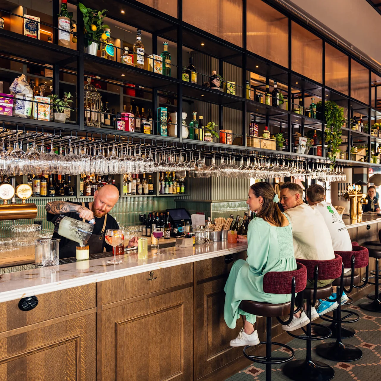 Bartender pours drinks for three patrons at a bar. Shelves filled with bottles and plants are visible above, in a brightly lit, modern setting.