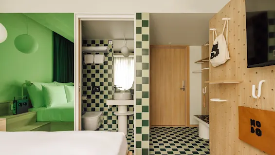 A modern hotel room featuring a vibrant green sleeping area, a checkered bathroom, and a wooden pegboard wall with a bag.