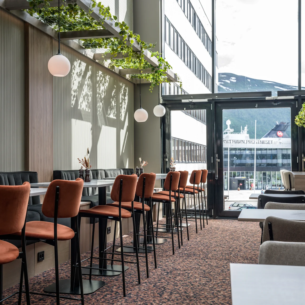Dining tables and orange chairs stand in a sunlit restaurant with pendant lights and greenery, adjacent to large windows showing a cityscape. Visible text: "IS7HAVN PRO LINESET."