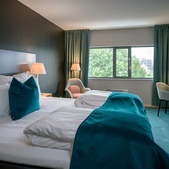 En pent oppredd seng med turkise innslag står i et mykt opplyst hotellrom, flankert av en stol og skrivebord. Store vinduer viser grønt utenfor.