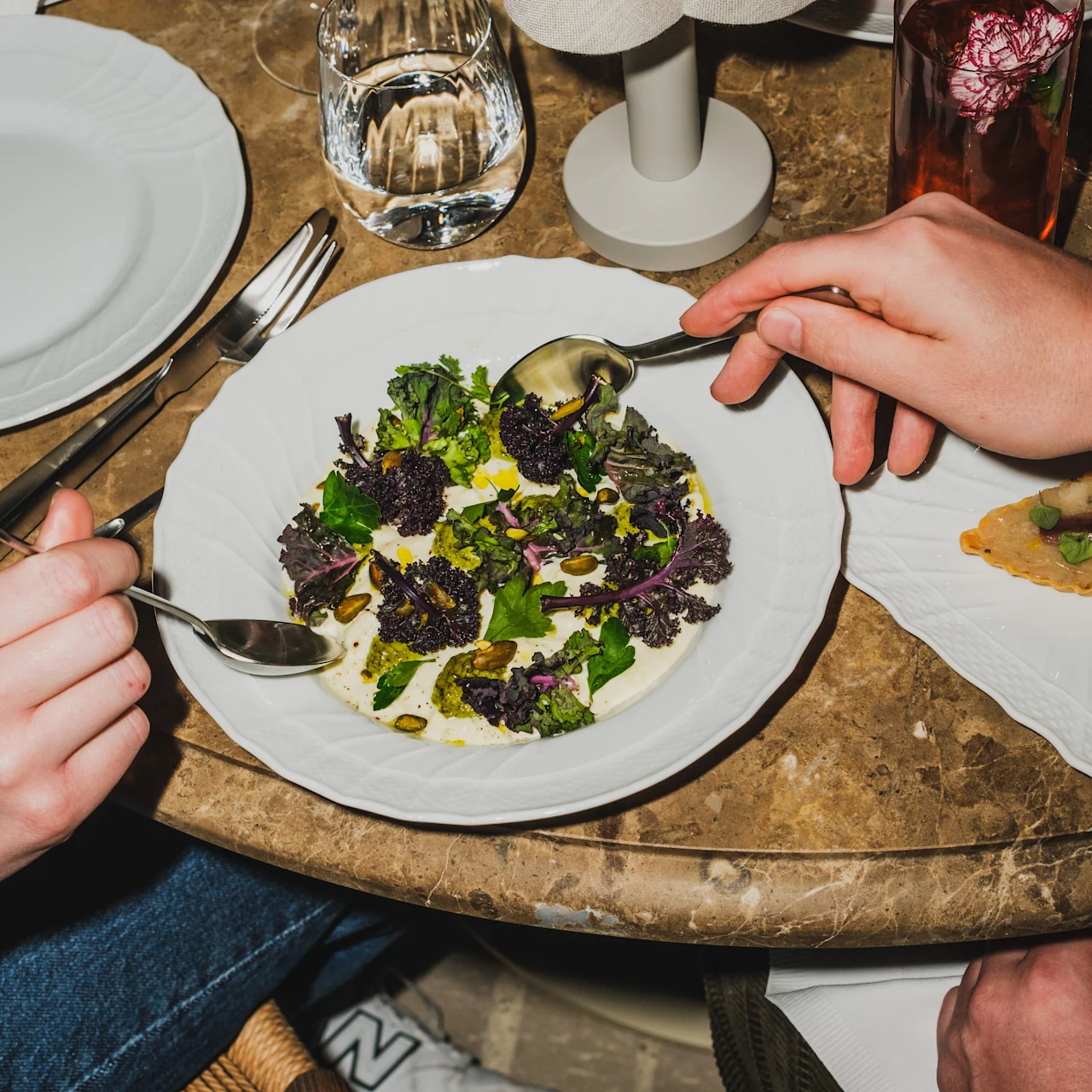 Two hands hold spoons over a salad with greens and seeds on a white plate. They sit at a marble table with utensils, a water glass, and a decorative flower.