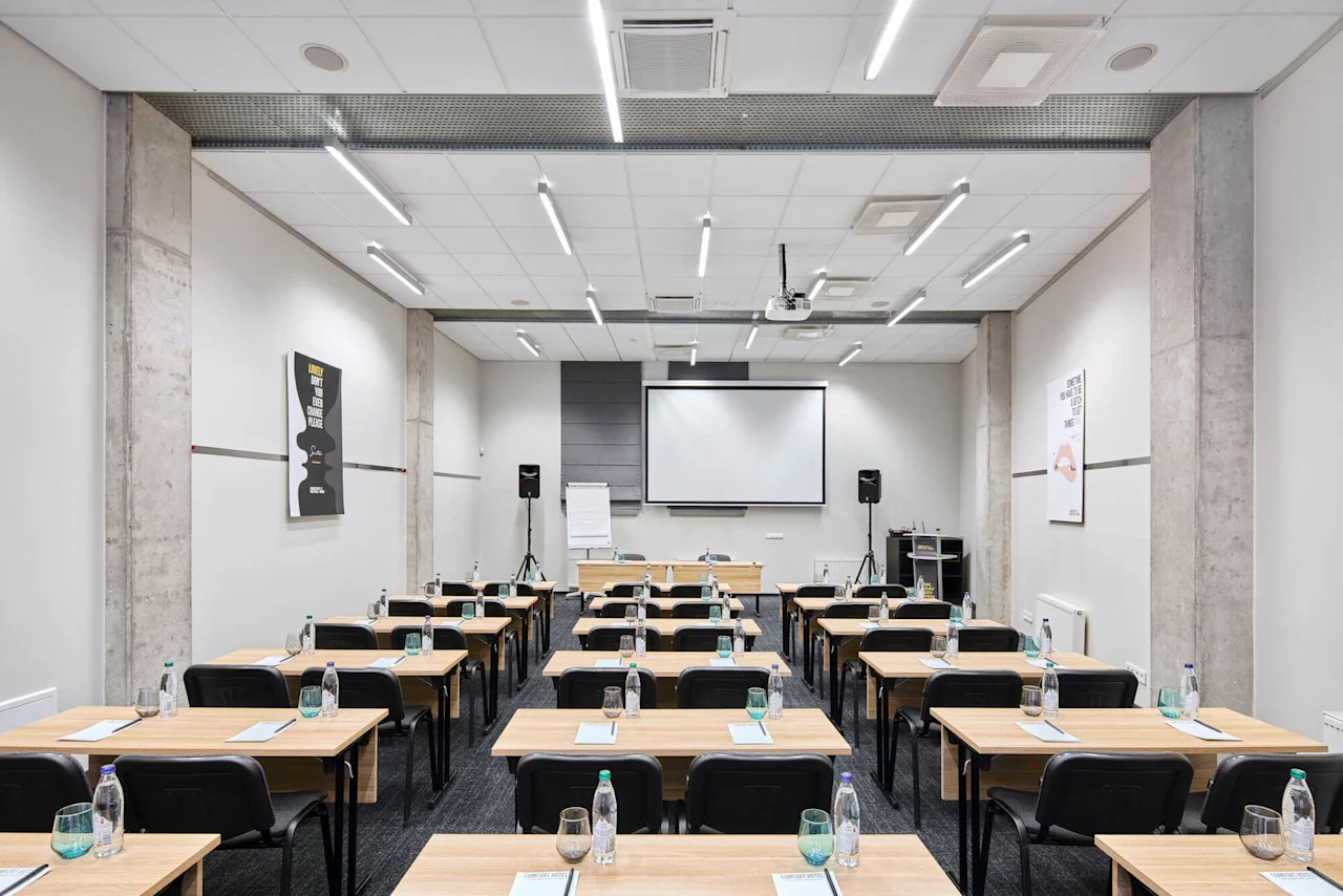 Classroom setup with wooden desks, black chairs, and bottled water. A projector screen hangs at the front, surrounded by speakers. Concrete walls display minimal posters.