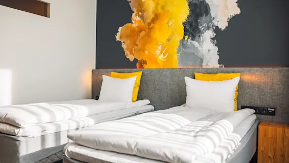 Two neatly made single beds with white linens and yellow accents, positioned against a wall featuring a vibrant abstract mural, in a brightly lit modern room. 