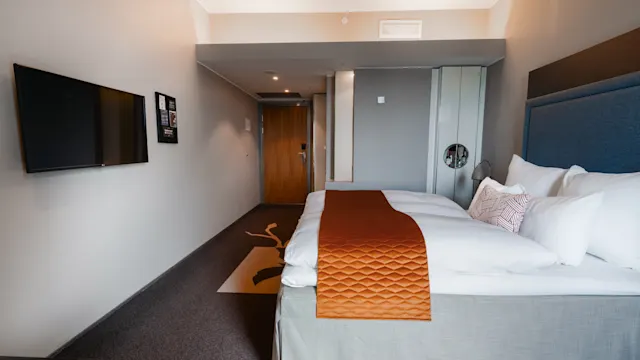 Siististi pedattu sänky valkoisilla liinavaatteilla ja oranssilla huovalla sijaitsee modernissa hotellihuoneessa. Seinälle kiinnitetty televisio on sänkyä vastapäätä, taustalla puuovi.