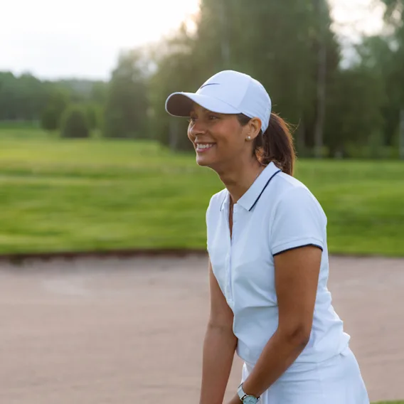 En person i hvit caps og pikétrøye smiler mens de nyter en golfrunde på en solrik dag.