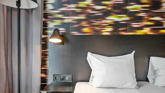 A bed with white pillows lies under a modern wall light, next to a colorful abstract patterned wall. Nearby, a floor lamp stands beside gray curtains.