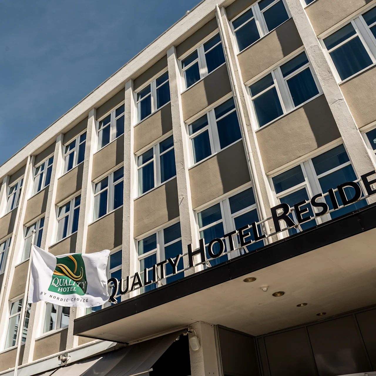 Nykyaikaisen monikerroksisen rakennuksen julkisivu, jossa on "QUALITY HOTEL RESIDENCE" -kyltti, monia ikkunoita ja lippuja. Yhdessä lipussa lukee "Quality Hotel by Nordic Choice". Kirkas, sininen taustataivas.