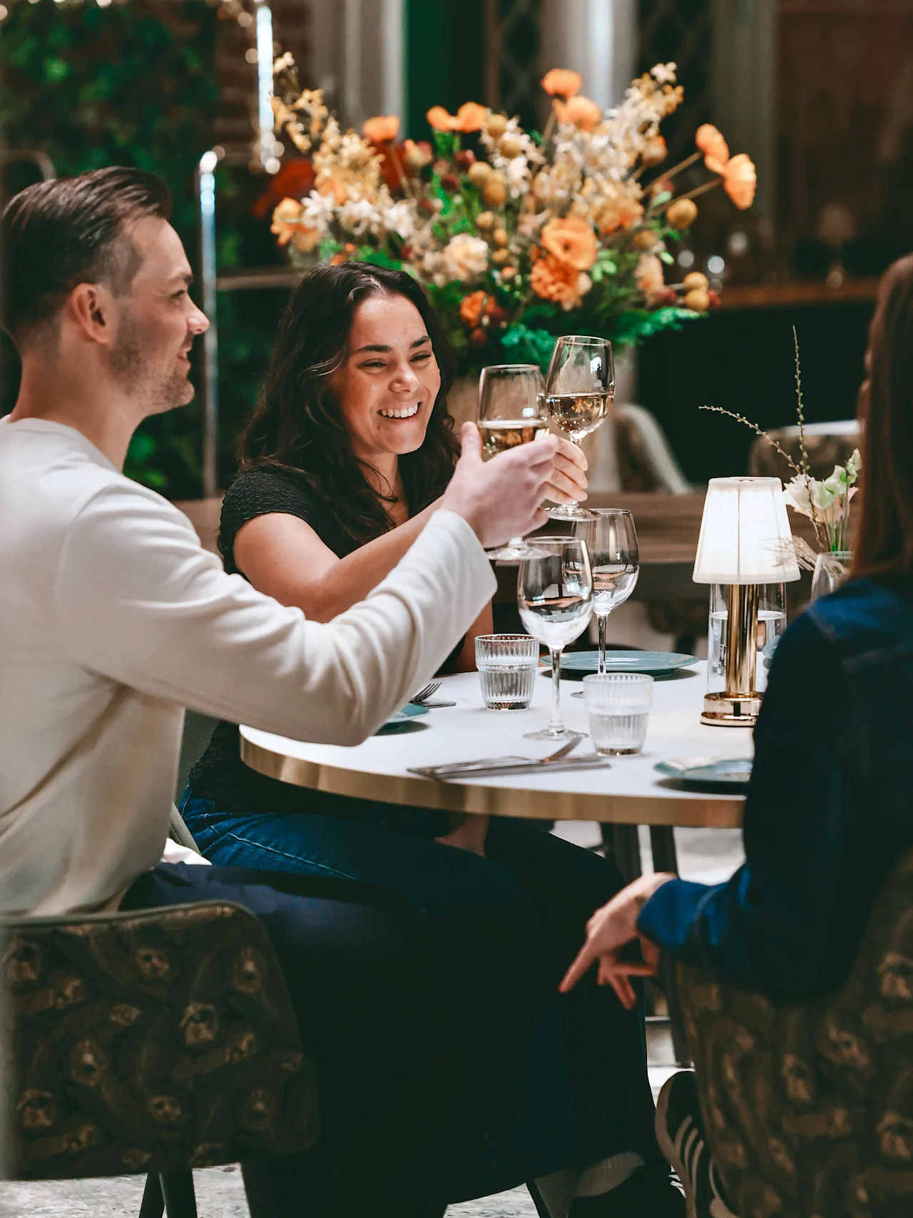 People at a table clinking wine glasses, smiling and laughing, with a floral centerpiece. Enjoying a social gathering.