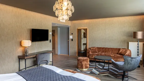 En seng med et gråt teppe står i et moderne rom med en lysekrone, veggmontert TV, bord med lampe, fløyelssofa, lenestol og geometrisk teppe på tregulv.