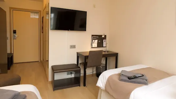 Et hotelværelse med en redt seng, sammenfoldede håndklæder og en fjernbetjening ovenpå, et vægmonteret TV og et skrivebord nedenunder. En lukket dør fører udenfor.