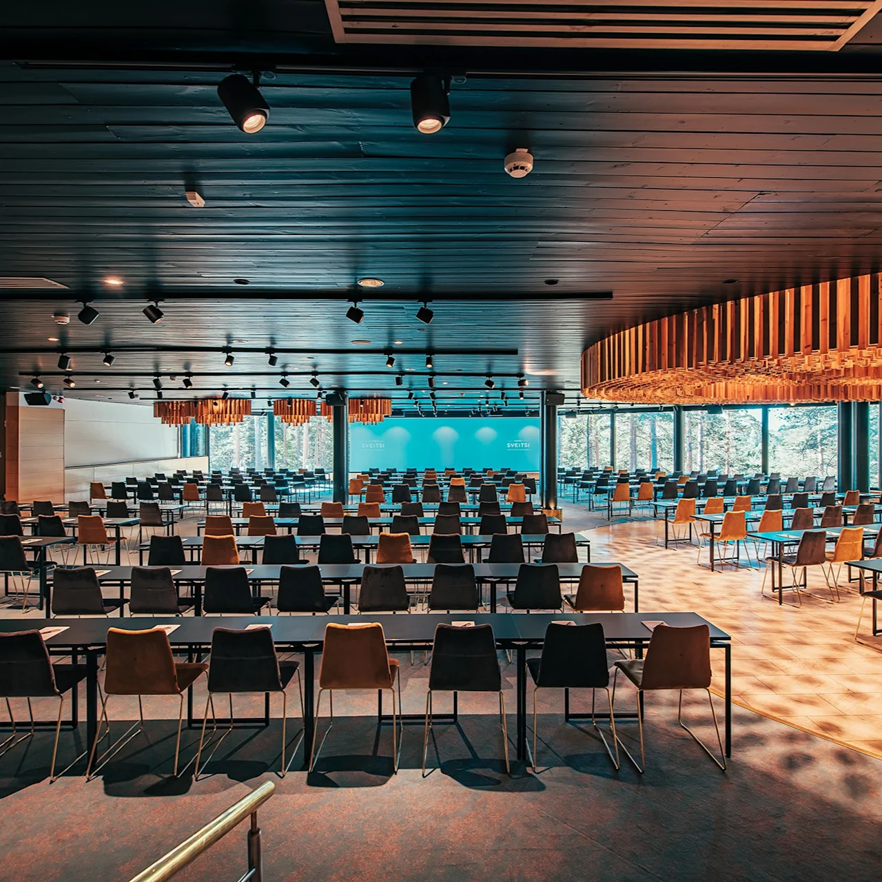 An empty conference room, with rows of chairs and rectangular tables, arranged neatly. Warm overhead lighting accentuates wooden features, while large windows offer a view of trees outside.
