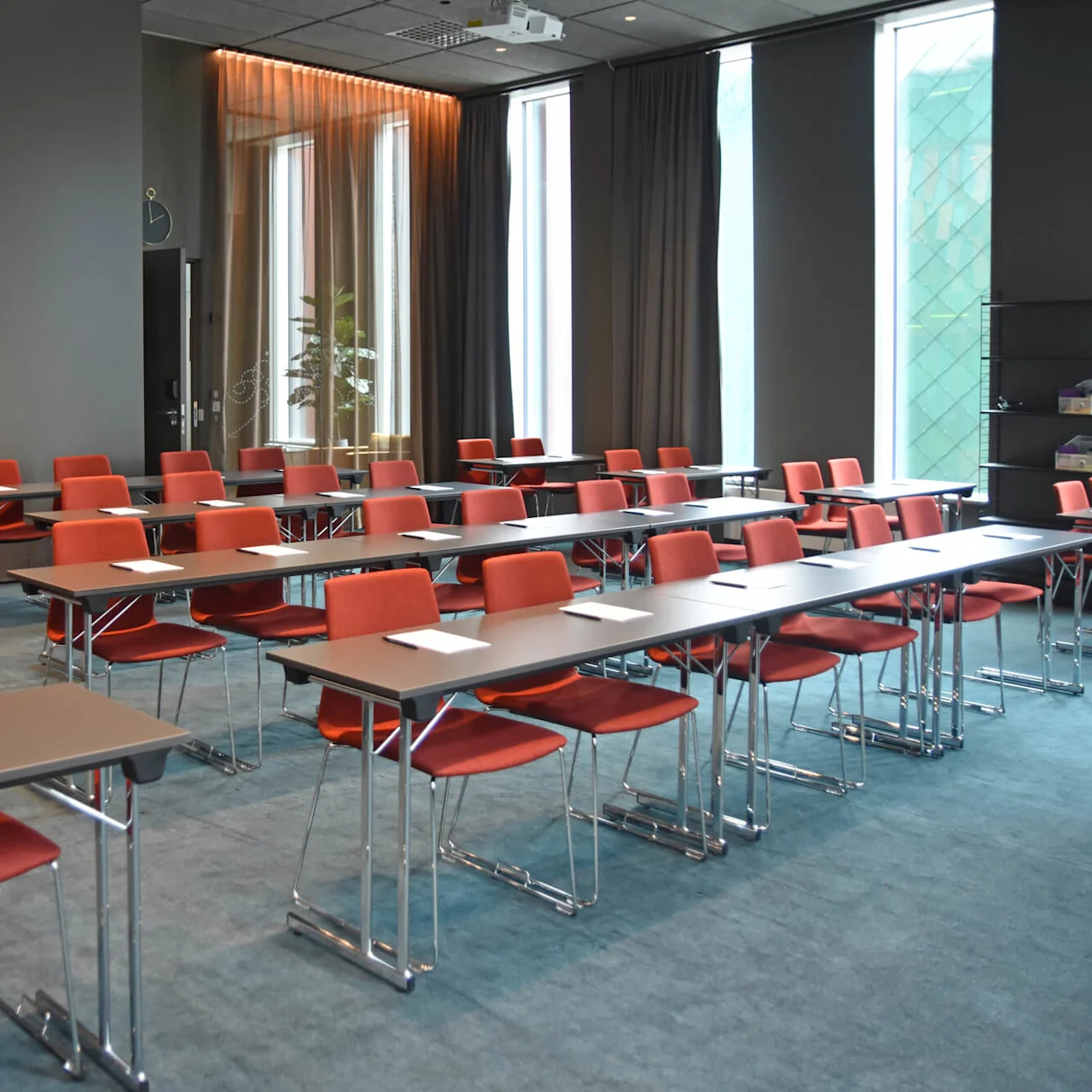 Rader av röda stolar och bord arrangerade i ett modernt konferensrum, med anteckningsblock och pennor på varje bord, omgivna av stora fönster och grå väggar med en klocka.