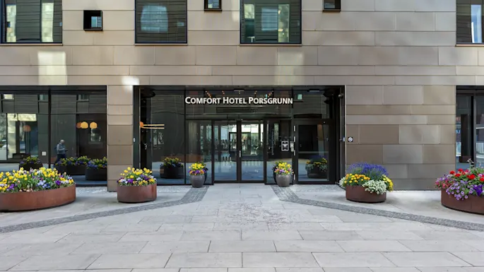 The entrance to Comfort Hotel Porsgrunn, featuring glass doors, a modern facade, and colorful flower planters.