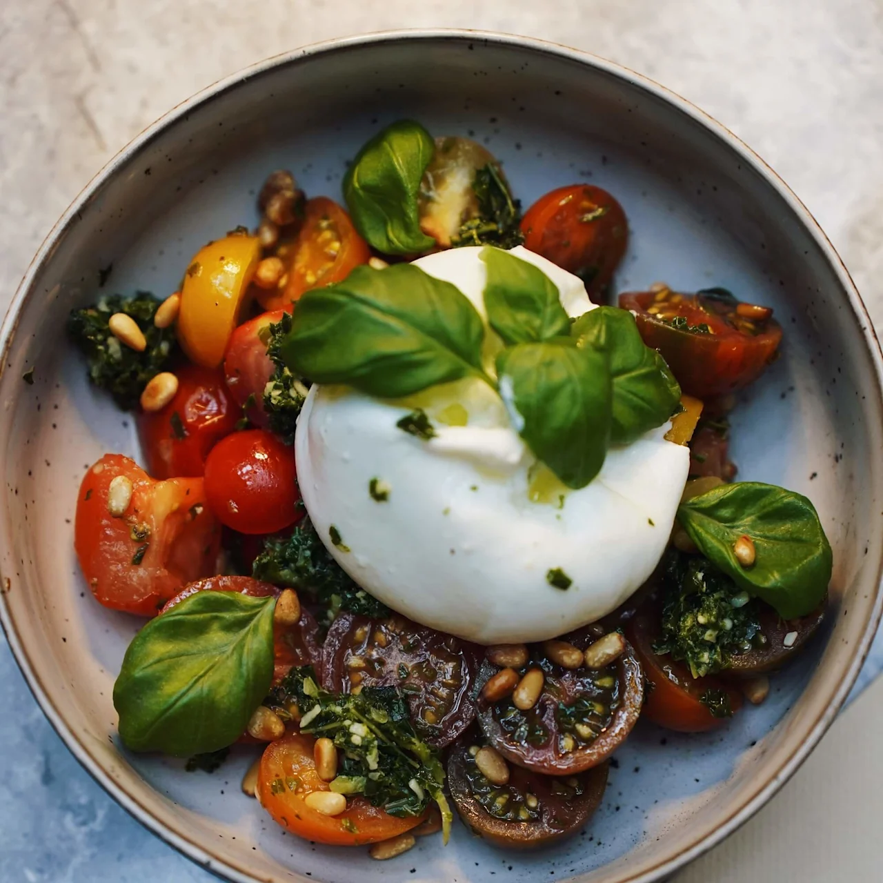Burrataost toppet med basilikum hviler på en seng av cherrytomater, pinjekerner og pesto. Anbragt i en plettet skål på et marmorbord, ved siden af et delvist synligt menukort med teksten "THE SOC".