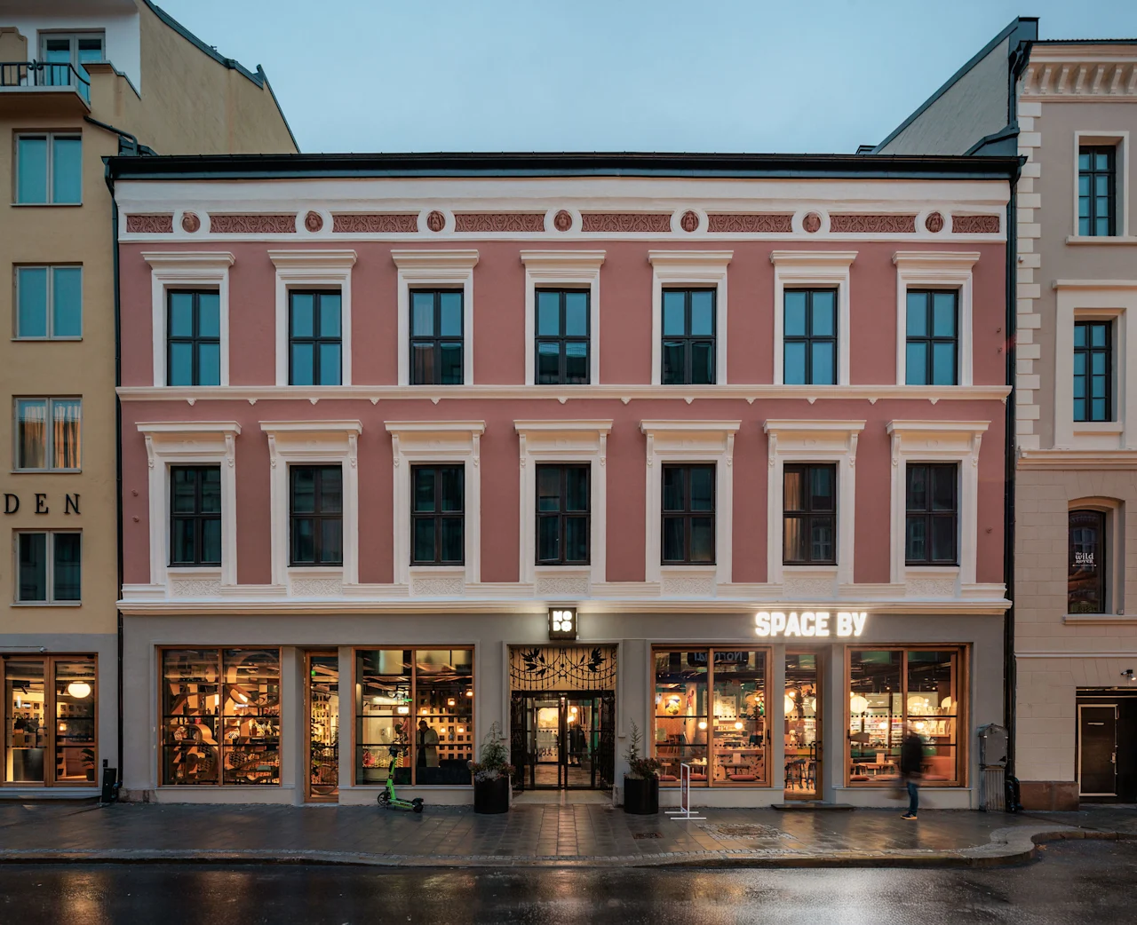 A pink building with white trim and many windows. The ground floor has large illuminated display windows and an entrance. Text reads SPACE BY.