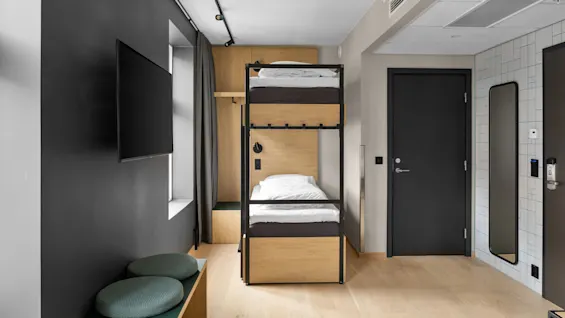 Våningssäng står i ett modernt sovrum; rummet har en väggmonterad TV, två gröna kuddar på en bänk, en spegel bredvid en svart dörr och en stilren, minimalistisk design.
