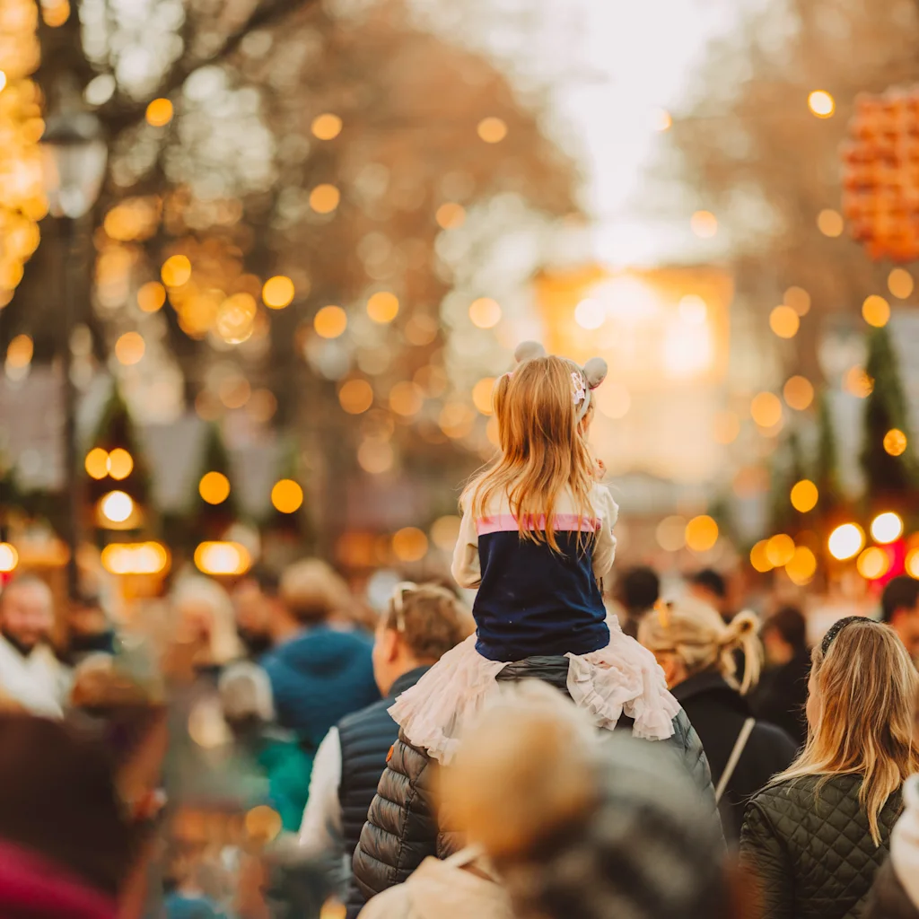 A child on someone's shoulders looking at a festive outdoor market with many lights and people.