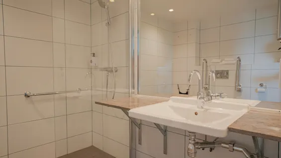 Et moderne badeværelse har en hvid vask med elegante armaturer på en bordplade, ved siden af en brusekabine med glasvægge. Rummet er omgivet af hvide flisevægge og indbygget lys.