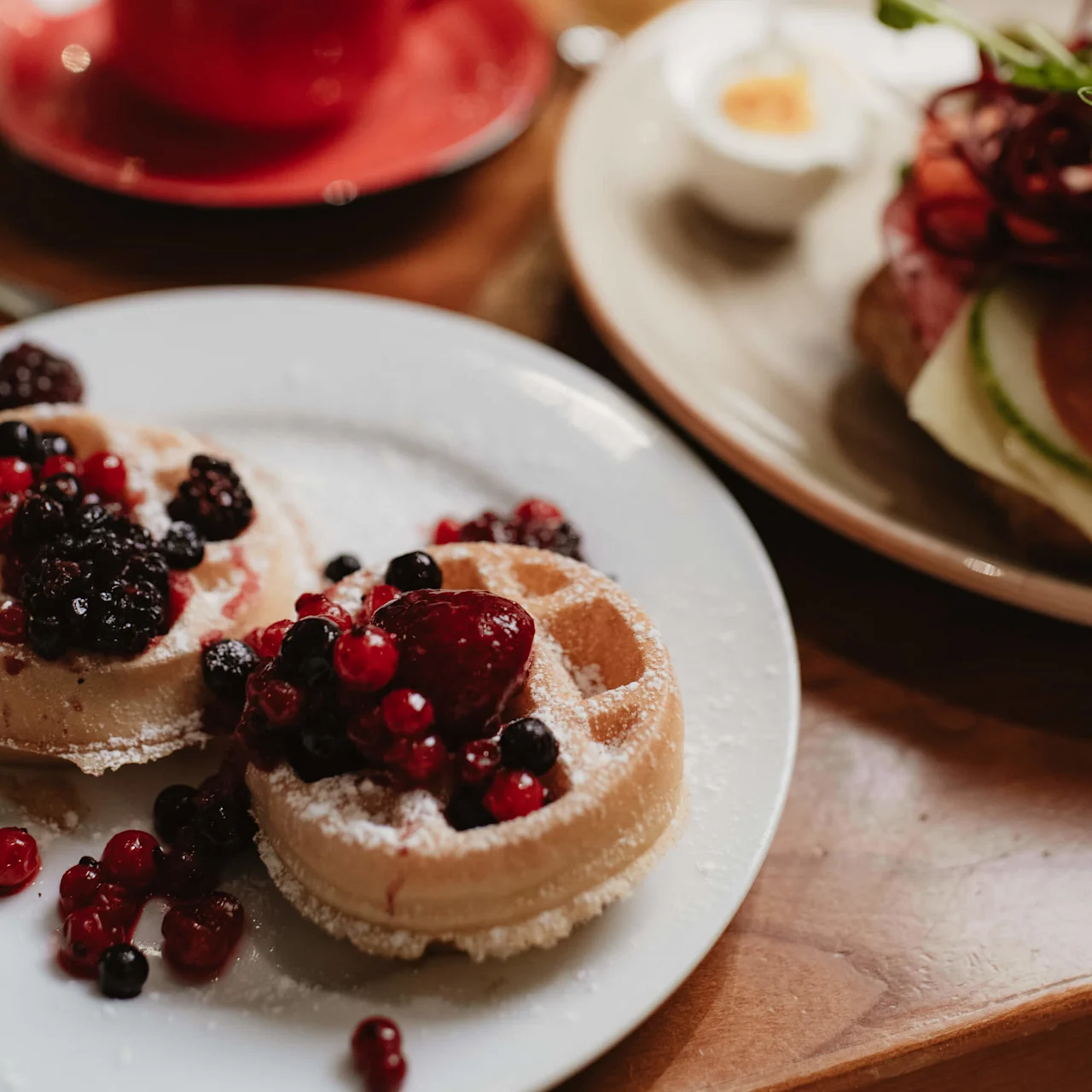 Waffles topped with berries and powdered sugar sit on a white plate. Nearby, a red coffee cup and a plate with a boiled egg and sandwich are on a wooden table.