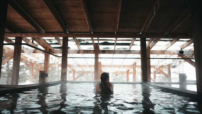 A person relaxes in a large indoor pool, surrounded by wooden beams and dim lighting, creating a cozy, serene atmosphere.