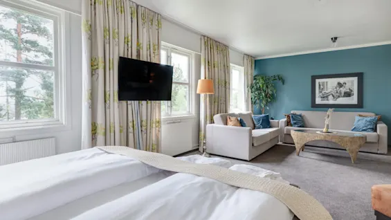 A bed with white linens faces a wall-mounted TV. Nearby, a cozy seating area includes a gray sofa with blue cushions and a wooden coffee table against a teal accent wall.