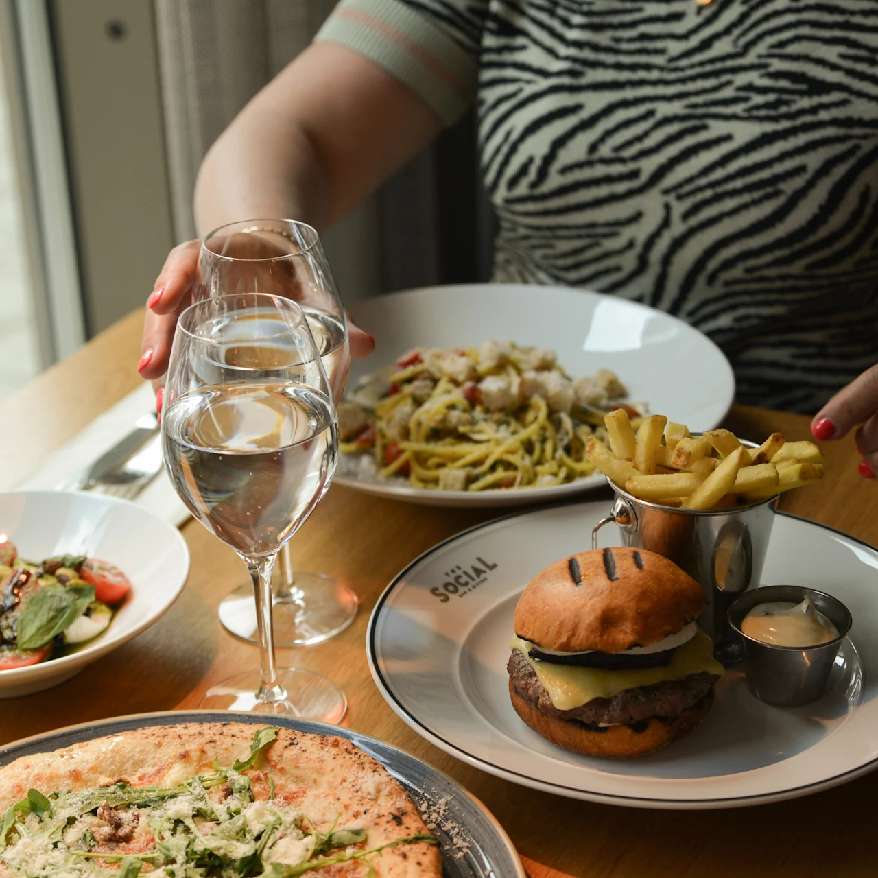 En person rækker ud efter et vinglas ved et bord med burger, pommes frites, pasta og salat serveret på tallerkener. Tekst på tallerkenen lyder "SOCIAL".