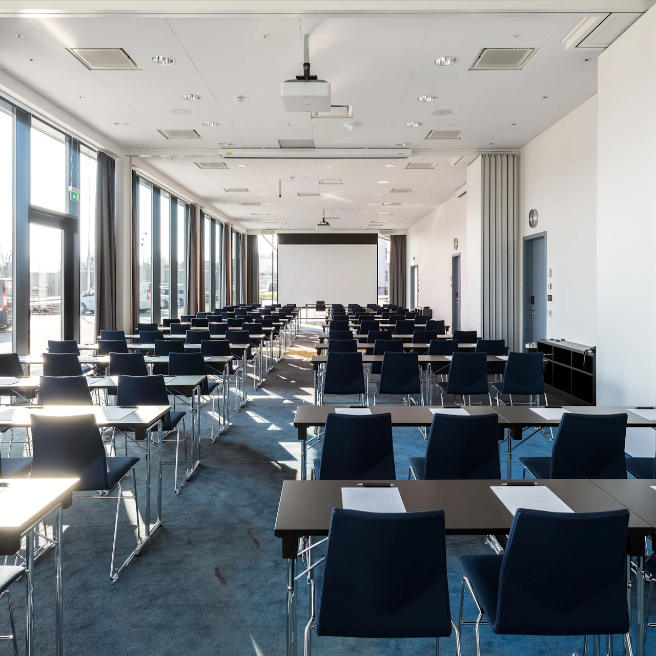 Rows of empty conference tables and chairs face a large projector screen in a spacious, sunlit room with tall windows and a blue carpeted floor.