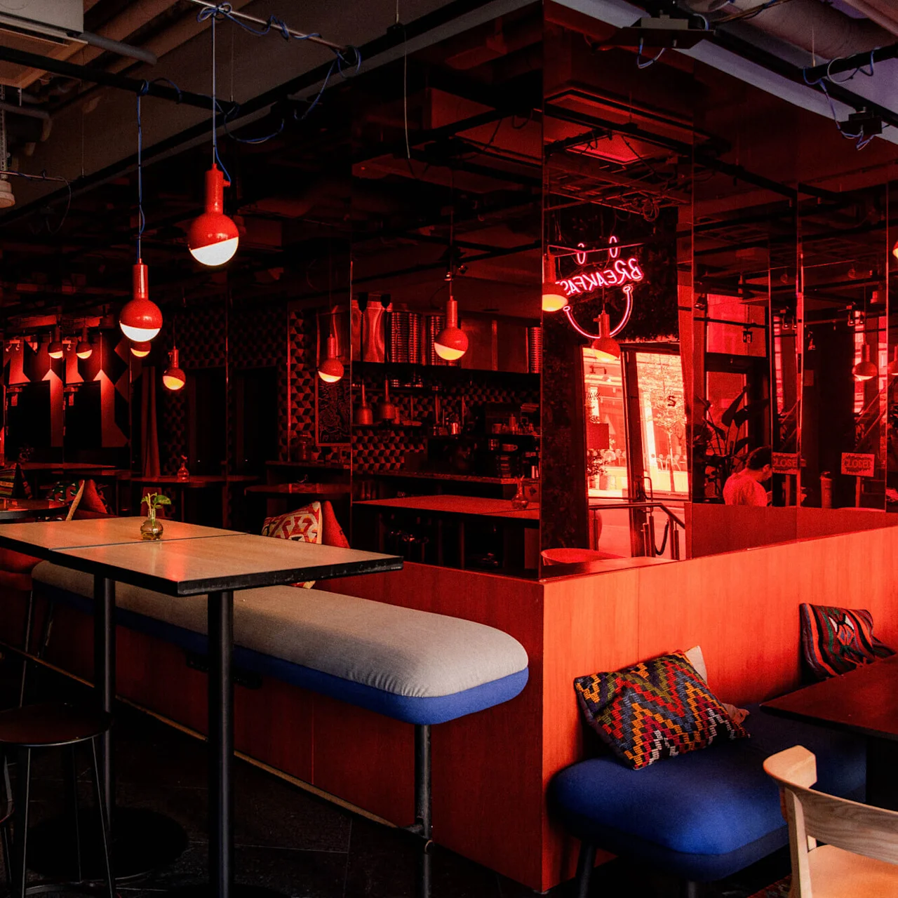 Red pendant lights illuminate tables and stools in a modern bar. A neon sign reads "BREAKFAST" and a smiley face. The bar is stylishly designed with geometric patterns.