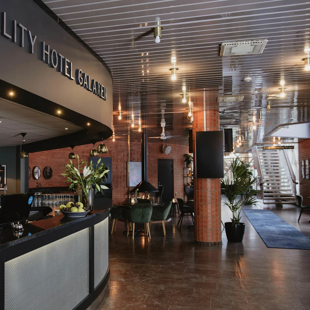 Reception desk displays "QUALITY HOTEL GALAXEN," with computers and snacks. Brick walls, green chairs, and tables in a warmly lit lobby. Stairs and plants enhance a modern feel.