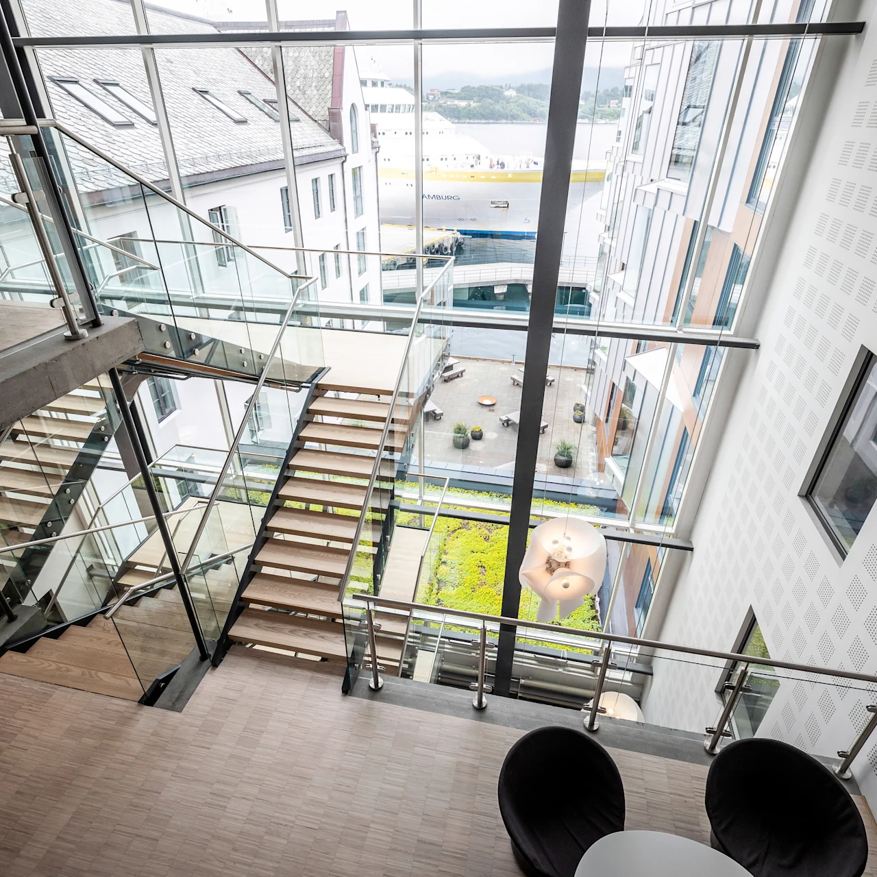 Trappan går ner i en modern, glasväggad byggnad med utsikt över en innergård och ett dockat skepp utanför. Två svarta stolar och ett bord syns nedanför.