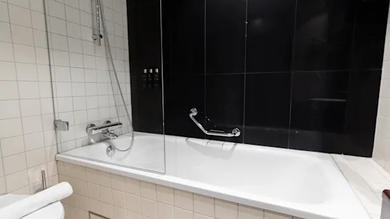 A white bathtub with a mounted showerhead is set against a dark tiled wall in a modern bathroom. A glass partition and towel on a toilet complete the design.
