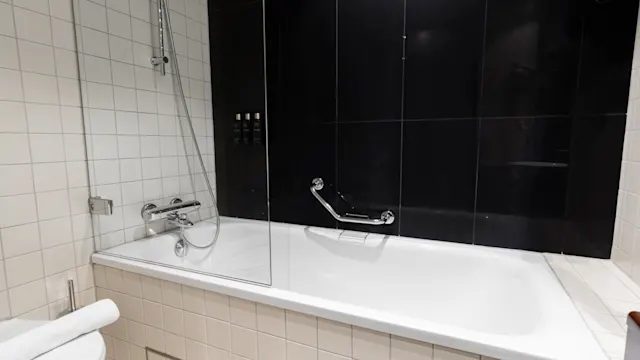 Valkoinen kylpyamme, jossa on asennettu suihku, on sijoitettu tummalle laatoitetulle seinälle moderniin kylpyhuoneeseen. Lasipaneeli ja pyyhe wc:ssä täydentävät muotoilun.