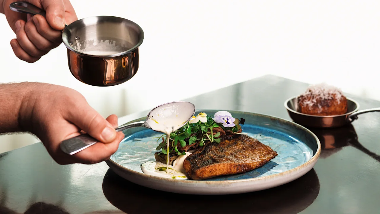 Hotel Kämp – Ravintola À La Kämp. Kokin kädet kaatavat varovasti kermaista kastiketta täydellisesti asetellun annoksen päälle, mikä parantaa esillepanoa.