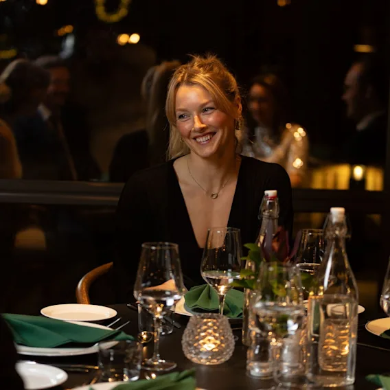 En lyshåret kvinne smiler ved et middagsbord i en restaurant dekket med vinglass og stearinlys.