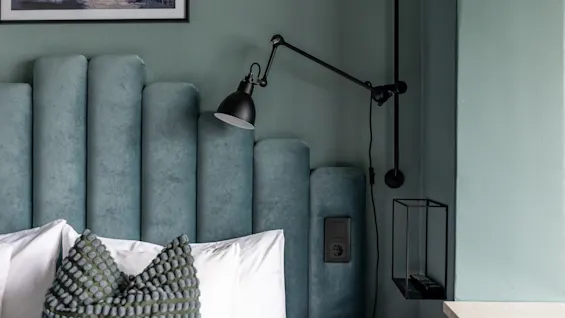 En komfortabel seng med en myk blå sengegavl, en moderne leselampe og en praktisk hylle for nødvendigheter.