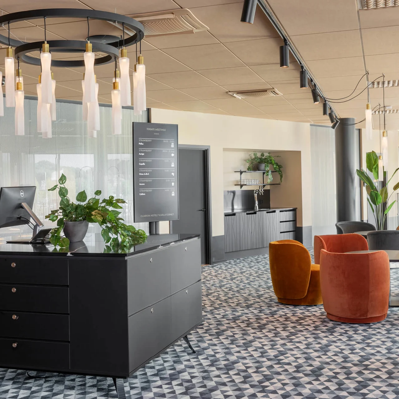Reception med dator och krukväxt, placerad i en modern lobby med geometriskt mönstrad matta, omgiven av orangea och grå stolar. Skylten lyder "DAGENS MÖTEN" med detaljerad schema.