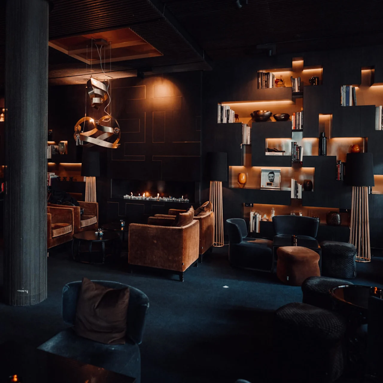 Luksuriøs lounge med moderne brune møbler, dempet belysning og en sentral peis. Hyller med bøker og dekor fyller veggene, noe som gir en varm, sofistikert atmosfære.